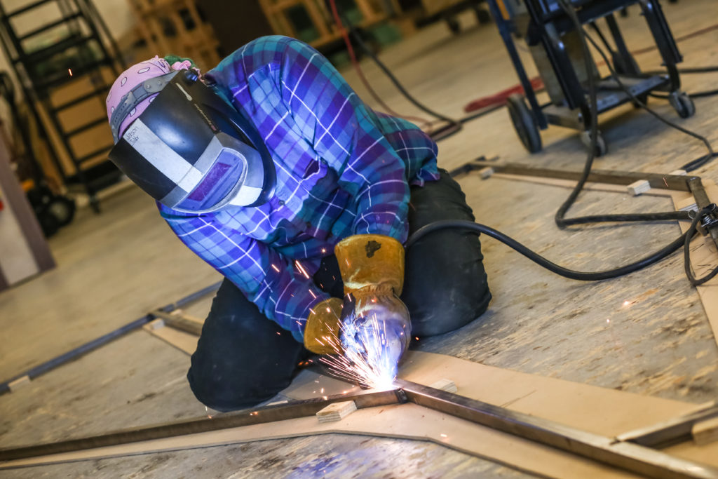 Sparks fly as a person in a flannel welds an angled steel frame that will become a tree trunk on stage.